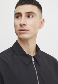 Sort lynlåsjakke med en moderat krave, prydet med en metal lynlås og en glat tekstur, båret af en model med kort hår.