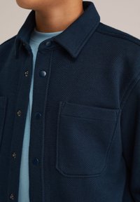 Pogled od blizu na osebo, ki nosi temno modro teksturirano jakno s gumbi čez svetlo modro majico, vidni so ovratnik in žep na prsih.