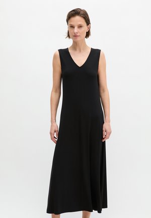 Femme debout portant une longue robe noire sans manches à col en V, sur un fond blanc uni.