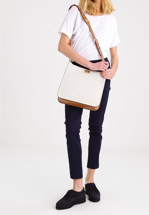 Femme portant une chemise blanche et un pantalon bleu marine, tenant un sac bandoulière blanc et marron avec un motif de logo, se tenant devant un fond uni.
