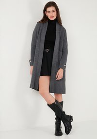 Gilet gris à côtes avec des manches larges, devant ouvert et poches, associé à un col roulé noir et une jupe, complété par des bottes noires montantes.