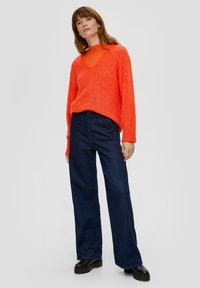 Felle oranje oversized trui met een v-hals, gecombineerd met donkerblauwe wijde jeans en zwarte platform schoenen. Gladde textuur, casual pasvorm.
