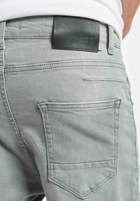 Jasnoszare dżinsy z denimu o gładkiej fakturze, z naszywką z skóry na pasku z napisem „DEF” oraz dwoma tylnymi kieszeniami ozdobionymi przeszyciami.