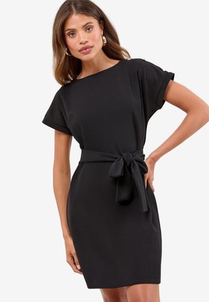 Vrouw die een kortmouwen zwarte jurk draagt met een gestrikte tailleband en gouden knoopdetails op de schouders, poseert met één hand op haar heup.