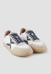 Baskets blanches avec des accents bleu marine et gris, comportant une finition en daim, un bout rond, une semelle épaisse en gomme et des lacets bleu marine. Logo de la marque sur la languette.