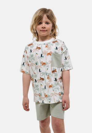 Enfant aux cheveux blonds mi-longs porte un t-shirt à manches courtes avec des animaux de la jungle et des palmiers, ainsi que des shorts vert clair.