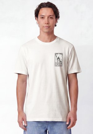 Homme aux cheveux courts et foncés portant un t-shirt blanc avec un petit motif noir de carte de tarot étiqueté "The Magician" sur la poitrine gauche.