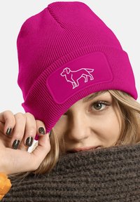 Magenta knit beanie with a ribbed design and a rectangular patch featuring a white dog silhouette. Soft texture and snug fit.