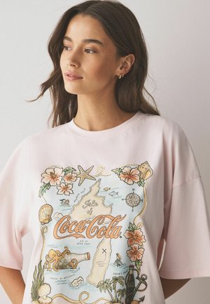 Mladá žena s dlhými tmavými vlasmi nosí svetloružové tričko Coca-Cola s grafikou tropickej mapy a morských tvorov.
