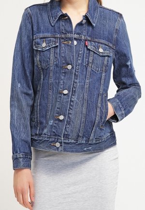 Vrouw met een blauwe spijkerjasje met knoopsluiting en voorzakken over een lichtgrijze rok, hand in het jaszak.