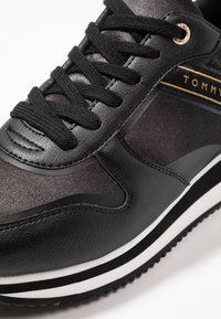 Černé tenisky s texturovaným svrškem z kůže a textilu, s černými tkaničkami, zlatými akcenty a bílou pruhovanou podešví.