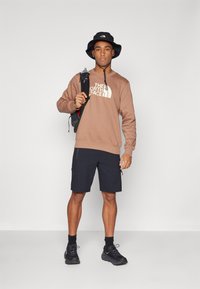 Felpa marrone con logo, pantaloni corti neri, cappello a secchiello nero e sneakers nere. Modello che tiene uno zaino, in piedi su uno sfondo neutro.