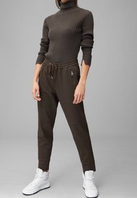 Brauner gerippter Rollkragenpullover kombiniert mit dunkelbraunen, taillierten Jogginghosen mit Kordelzug und Reißverschlusstaschen. Getragen mit weißen Sneakern.