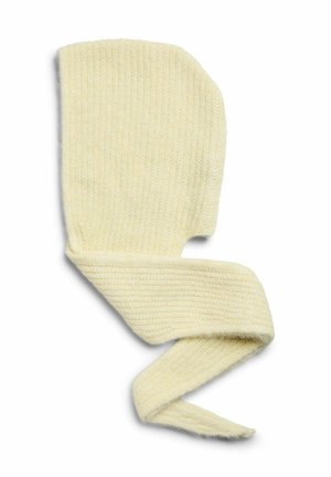 Una mujer luce en un estudio un abrigo largo de color antracita, una camiseta gris, unos pantalones oscuros holgados y una bufanda verde pálido envuelta alrededor de la cabeza.