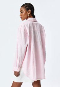 Pink long-sleeve shirt with a loose fit, featuring white heart patterns and a button-down collar. Soft, textured fabric with rolled cuffs.