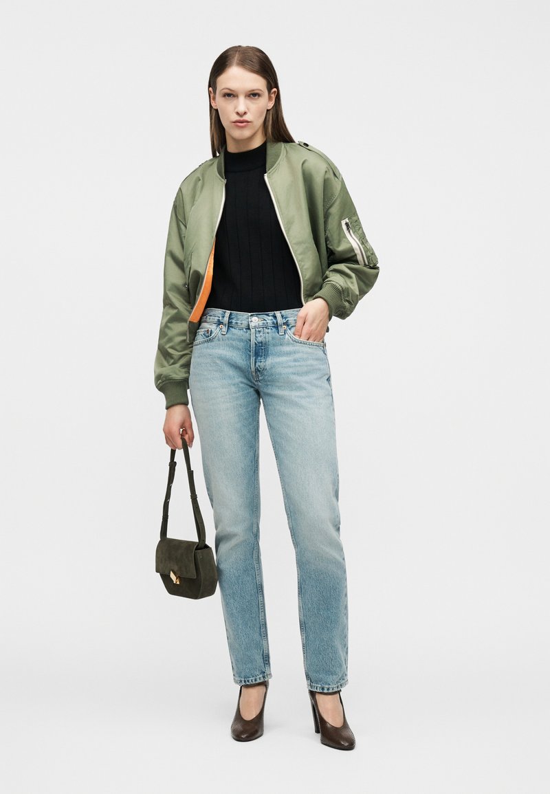 Femme portant une veste bomber verte, un haut noir, un jean bleu clair, des talons marron, tenant un petit sac à main vert foncé, debout devant un fond blanc.