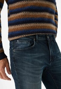 Uomo che indossa un maglione a righe blu, marrone e beige e jeans di denim blu scuro, con la mano rilassata al fianco, su sfondo bianco.