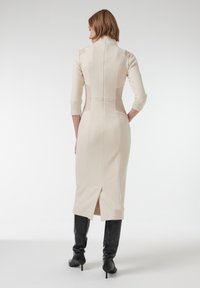 Beige aansluitend jurkje met een hoge kraag, driekwart mouwen en een split aan de achterkant, gemaakt van textiel met structuur. Gemaakt in combinatie met kniehoge zwarte laarzen.