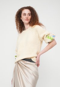 Femme aux cheveux roux bouclés portant un pull en maille crème avec une broderie florale sur les manches et une jupe en satin beige, posant devant un fond blanc.