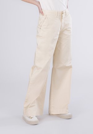 Kvinde iført løstsiddende beige bukser og hvide sneakers, stående med den ene hånd på hoften mod en ensfarvet baggrund.