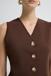 Canotta marrone senza maniche con scollatura a V e tre irregolari bottoni dorati lucidi, indossata da una persona con orecchini a cerchio dorati.