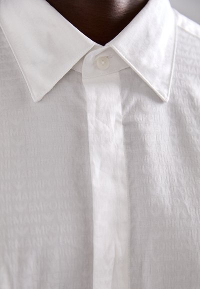 Balta pogu krekls ir izgatavots no teksturēta auduma ar smalku Emporio Armani logo rakstu. Tam ir klasiskā apkakle un viena pogu aizdares.