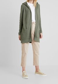 Grøn strikket cardigan med hætte, der har to frontlommer, båret over en hvid t-shirt og parret med beigesfarvede cropped bukser og beige sneakers.