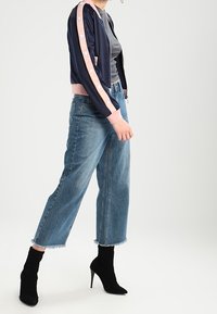 Kvinna i avklippta vida jeans, svarta högklackade stövlar, grå topp och marinblå bomberjacka med rosa rand och tryckknappar på ärmarna.