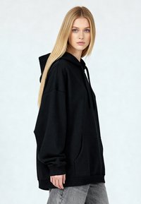 Črna oversized majica s kapuco z vrvico, sprednjim žepom, rebrastimi manšetami in gladko teksturo. Model nosi svetlo sive kavbojke.