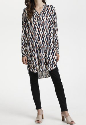Femme portant une tunique à manches longues, longueur genoux, avec des motifs géométriques bleu marine, blanc et marron, pantalon slim noir et chaussures à talons ouvertes beige.
