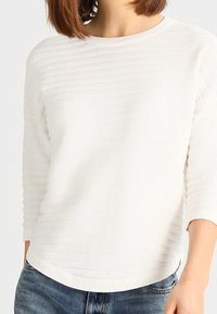 Haut blanc à manches longues avec des rayures horizontales en relief, un col rond et un ourlet légèrement courbé. Texture douce, coupe décontractée.