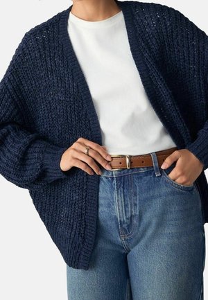 Personne portant un cardigan en grosse maille bleu marine au-dessus d'un t-shirt blanc, rentré dans un jean bleu taille haute avec une ceinture en cuir marron.