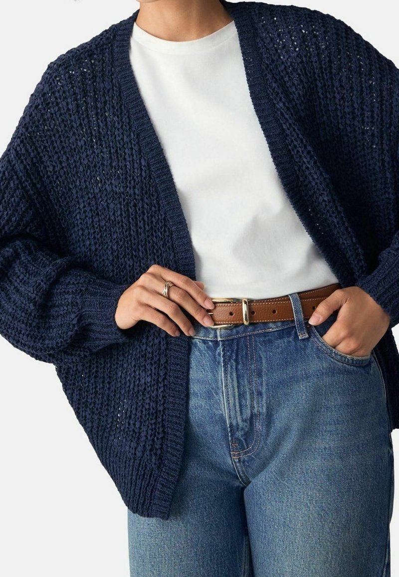 Personne portant un cardigan en grosse maille bleu marine au-dessus d'un t-shirt blanc, rentré dans un jean bleu taille haute avec une ceinture en cuir marron.