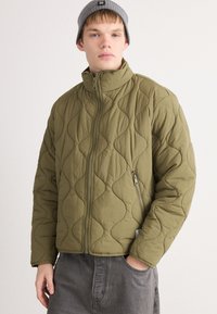 Joven vestido con una chaqueta acolchada verde olivo con cremallera y un gorro gris, con una mano en el bolsillo, de pie contra un fondo liso.