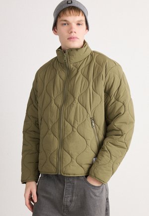 Joven vestido con una chaqueta acolchada verde olivo con cremallera y un gorro gris, con una mano en el bolsillo, de pie contra un fondo liso.