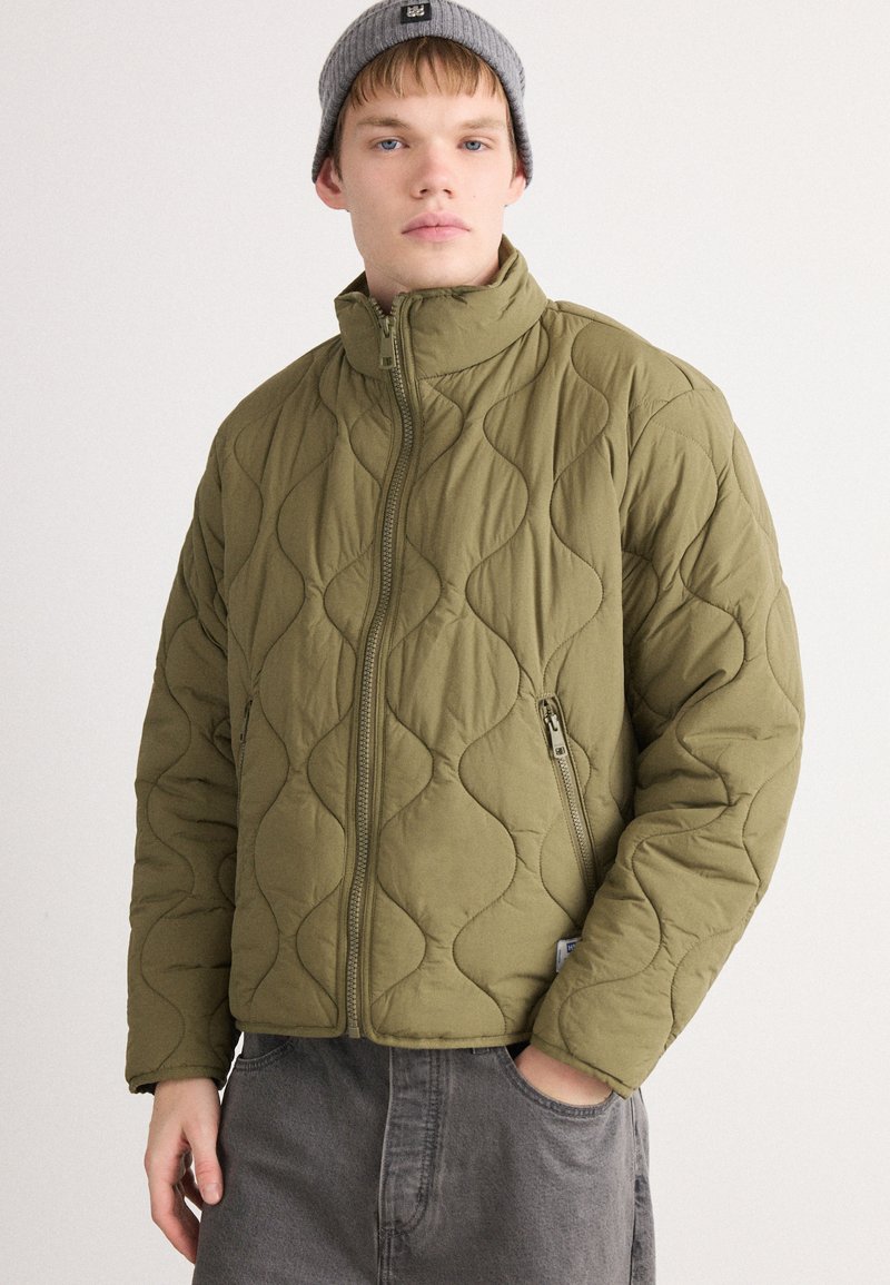 Joven vestido con una chaqueta acolchada verde olivo con cremallera y un gorro gris, con una mano en el bolsillo, de pie contra un fondo liso.