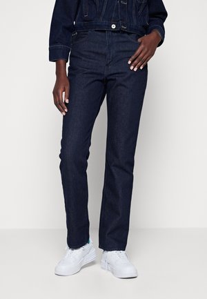 Personne portant un jean denim bleu foncé et une veste avec des baskets blanches, debout devant un fond clair uni.