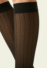 Piernas con pantimedias transparentes negras con diseños de diamantes y líneas verticales, mostradas desde la mitad del muslo hasta debajo de la rodilla.