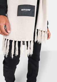 Beige gebreide sjaal met franjes aan de uiteinden, met een zwart rechthoekig label waar de merknaam "SIXTH JUNE" in het wit op is gedrukt.
