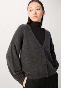Siv štrikan cardigan z V-izrezom, dolgimi rokavi in petimi gumbi spredaj. Tekstura se zdi mehka in udobna.