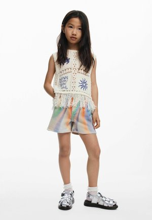 Háčkovaný top s strapcami a modrou výšivkou. Farebné šortky s efektom tie-dye. Strieborné sandále s remienkami a biele ponožky.