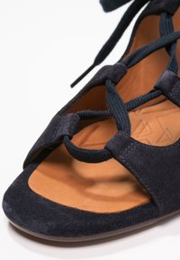 Sandale en daim marine avec bretelles croisées en corde et semelle intérieure en cuir marron. Design à bout ouvert avec une texture lisse et des accents métalliques minimalistes.