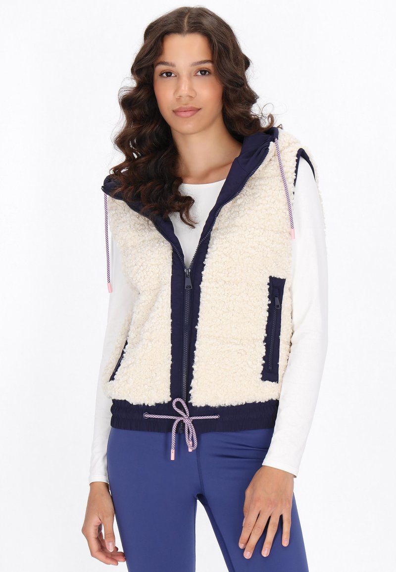Creme fuzzy vest met marineblauwe zijkanten, volledige rits, zakken, trekkoord onderrand en een capuchon. Textuurstof contrasteert met de gladde delen.