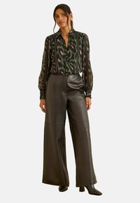 Nő zöld és lila mintás blúzban, ami bele van tűrve a magas derekú, bő szárú fekete bőrnadrágjába, fekete magassarkú csizmával.