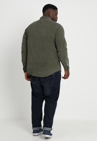 Camicia di velluto a coste verde oliva con maniche lunghe, orlo curvo e polsini con bottoni, abbinata a jeans in denim scuro a vestibilità rilassata.