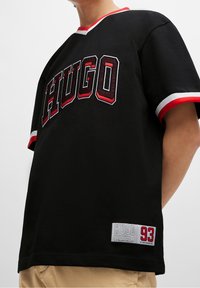 Svart bomull T-shirt med röda accenter, präglad med "HUGO" i fet stil, och en etikett längst ner med numret "93" och logotyp.