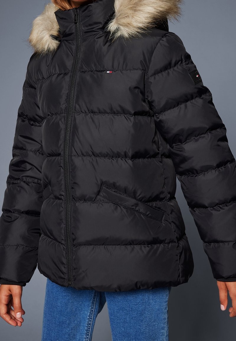 Veste matelassée noire avec capuche bordée de fourrure, fermeture éclair frontale, poches latérales et petits écussons logo sur la poitrine et la manche.
