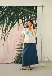 Femme en blouse blanche brodée, longue jupe en denim et sandales noires, debout à côté d'un arrière-plan avec de grandes feuilles de palmier vertes.