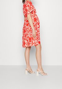 Röd blommig gravidklänning med korta ärmar och lös passform; mjukt tyg; matchad med vita blockklacksandaler.