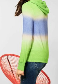 Grön och blå gradient hoodie med avslappnad passform, som har långa ärmar och dragsko i huvan. Bärs med blå jeans, röd stol i bakgrunden.